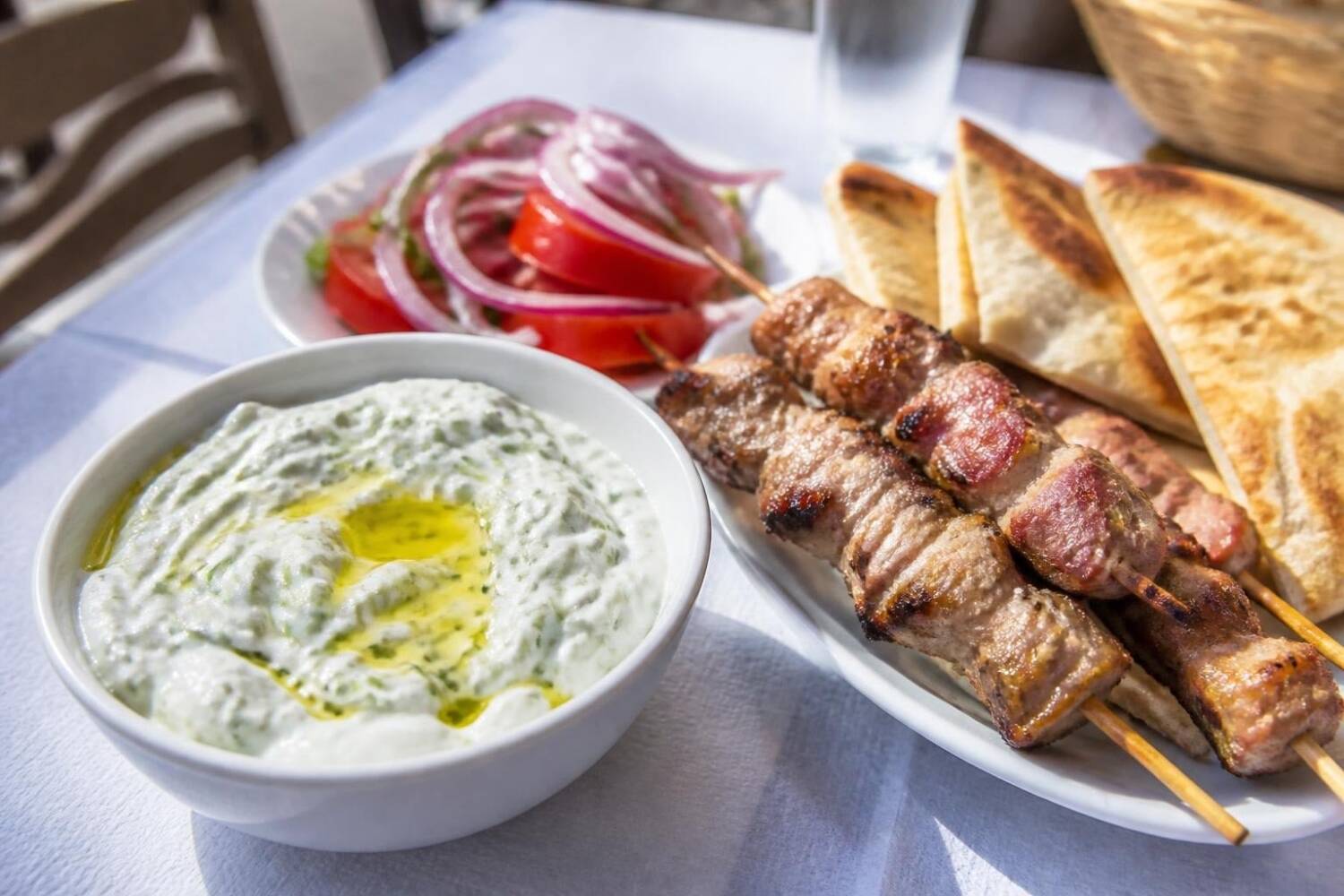 Tzatziki mit Olivenöl, Souvlaki-Spieße, Pita und Tomatensalat auf einem Tavernentisch
